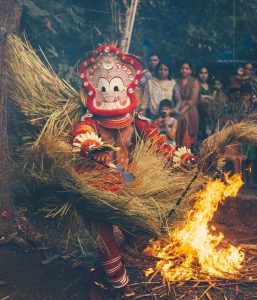 Pottan Teyyam surrounded by flames and torches moves through a nighttime ritual space, sparks filling the air.