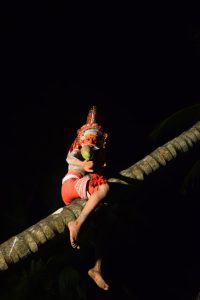 Bhappiriyan Teyyam sits on a coconut tree trunk at night, holding a coconut and lit dramatically against the dark sky.