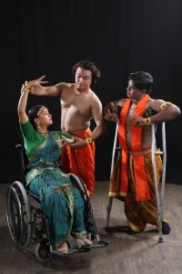 Three Bharatanatyam dancers, one using a wheelchair and one using crutches, alongside a standing nondisabled dancer. The standing Bharatanatyam dancer adjusts the arm positions of the wheelchair dancers, guiding their classical mudras.
