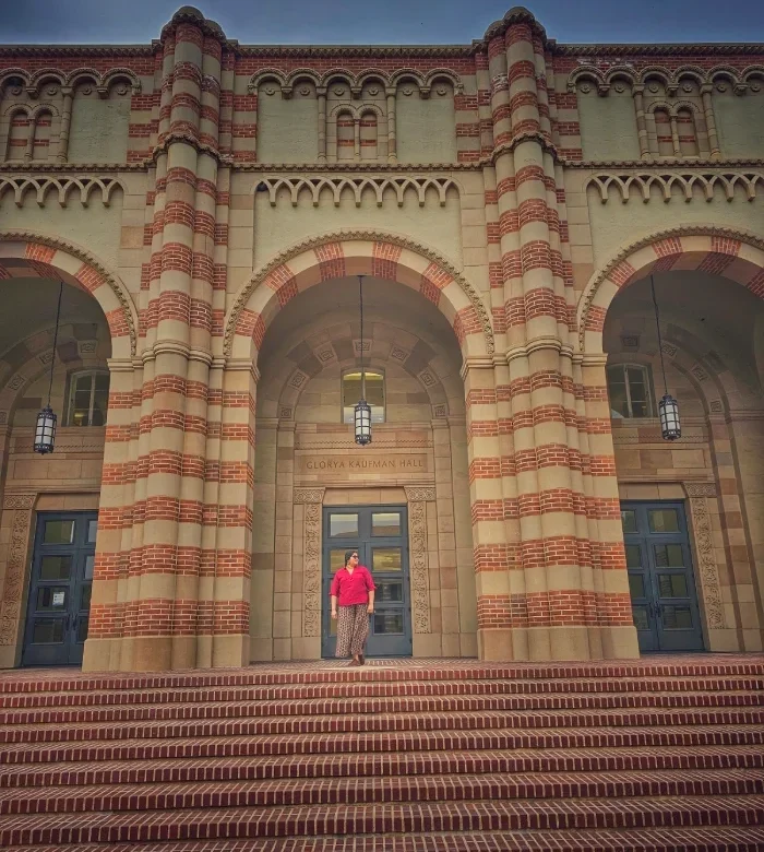 A person in a red top and patterned skirt stands centered on the wide brick steps of Glorney Kaufman Hall, framed by tall arched doorways and ornate brickwork.