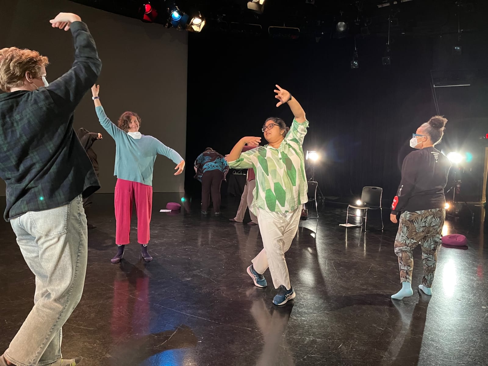 Several dancers stand in a studio lit by stage lights, each holding a different expressive pose. One person lifts an arm overhead, another leans into a sideways gesture, and others stretch or twist as they rehearse.
