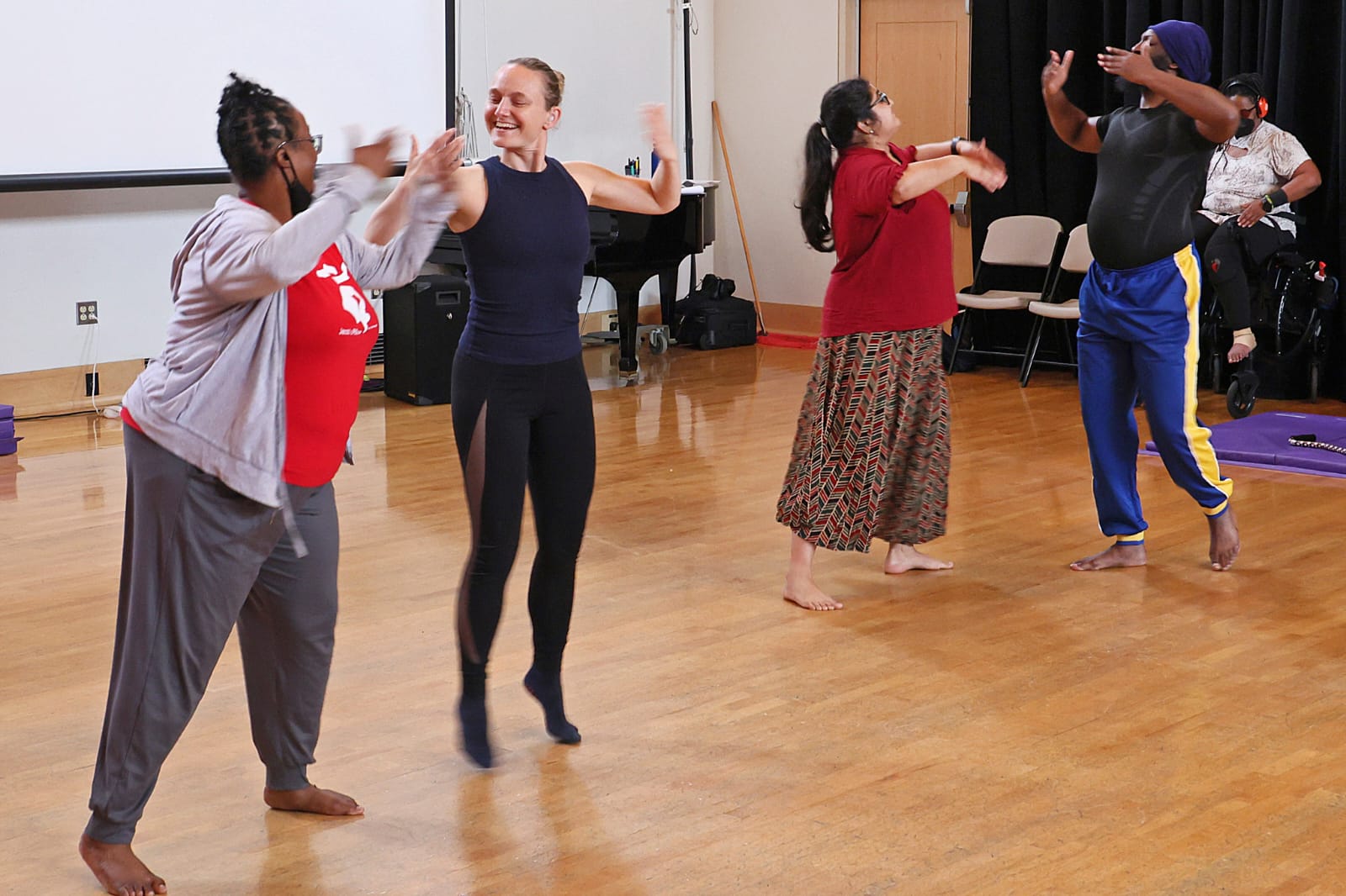 Four dancers share lifted gestures, meeting each other’s arms with a sense of ease and play. A dancer in a wheelchair watches from the background as the group moves across the studio floor, shaping a moment that blends movement, touch, and ASL-informed expression within a mixed Deaf and VI performance practice.