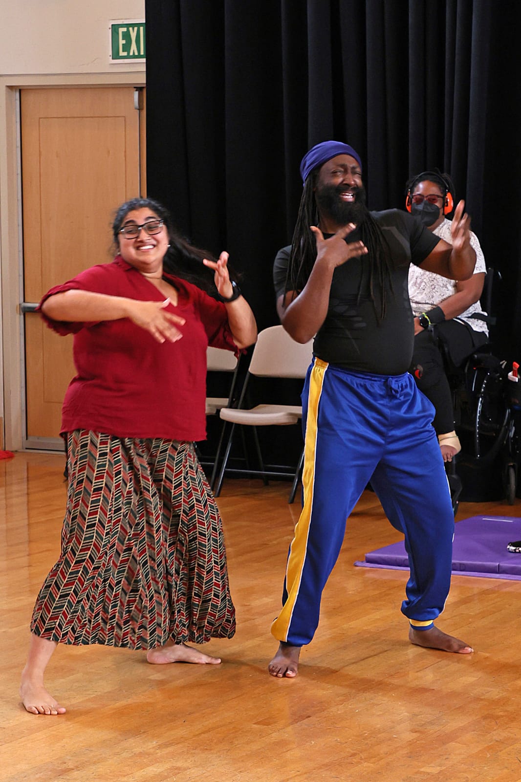 Two dancers move side by side, bodies loose and expressive, their hands carving gestures through the air while others watch from behind.