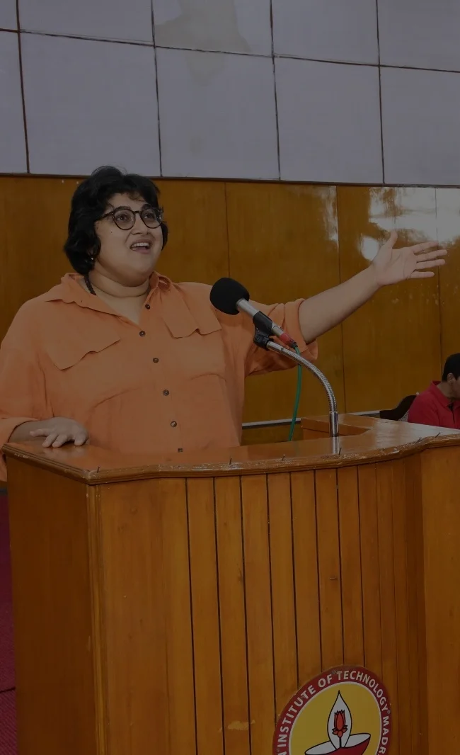 A woman stands behind a wooden lectern in a lecture hall, speaking into a fixed microphone. She wears an orange shirt and round eyeglasses, with short, slightly tousled dark hair. One arm extends outward in a broad, expressive gesture as she emphasizes a point, while her other hand rests on the lectern. Her mouth is open mid-speech, suggesting active engagement with the audience. Paneled walls and seated listeners are visible in the background, reinforcing the formal, public speaking setting.