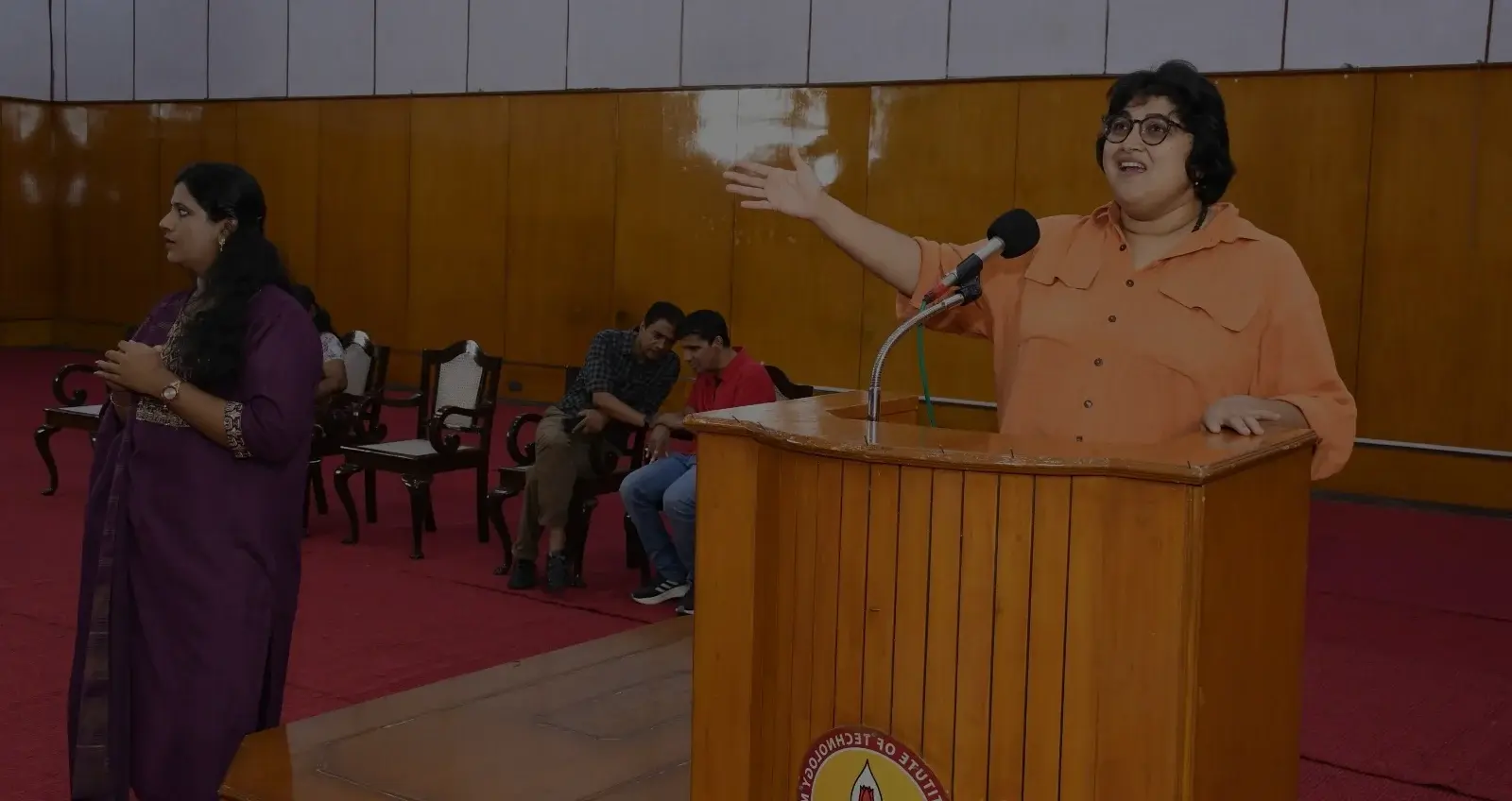 A woman stands behind a wooden lectern in a lecture hall, speaking into a fixed microphone. She wears an orange shirt and round eyeglasses, with short, slightly tousled dark hair. One arm extends outward in a broad, expressive gesture as she emphasizes a point, while her other hand rests on the lectern. Her mouth is open mid-speech, suggesting active engagement with the audience. Paneled walls and seated listeners are visible in the background, reinforcing the formal, public speaking setting.