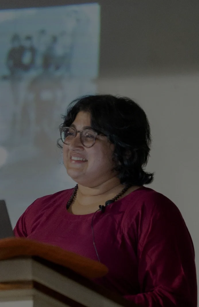 A woman stands indoors, shown from the waist up, wearing a deep maroon top and round eyeglasses. She has short, dark hair framing her face and a small microphone clipped near her collar, indicating she is presenting. The background is softly blurred, with a projected image visible on a screen behind her, situating her in an academic or lecture setting.