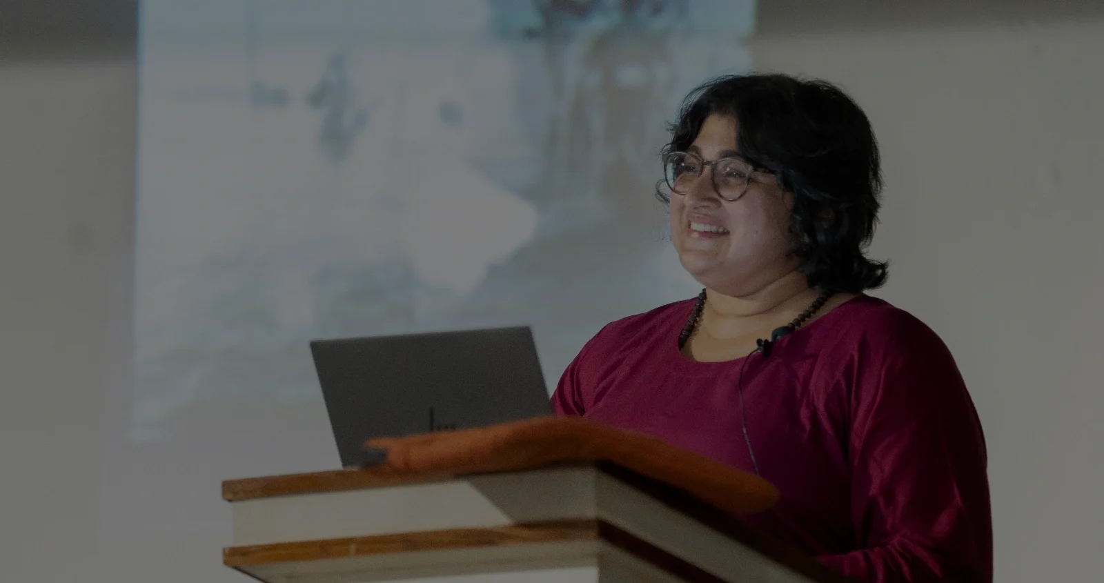 A woman stands indoors, shown from the waist up, wearing a deep maroon top and round eyeglasses. She has short, dark hair framing her face and a small microphone clipped near her collar, indicating she is presenting. The background is softly blurred, with a projected image visible on a screen behind her, situating her in an academic or lecture setting.